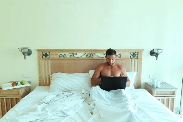 young naked man in bedroom laying in bed with laptop working at the morning. Handsome man needs to work on holiday to make money and be successful on his job. Guy with notebook is busy working at home
