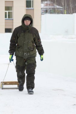 Sağ elinde kızak olan bir adam buzlu kasaba boyunca yürüyor.