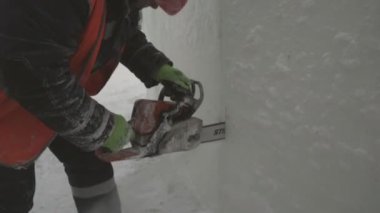 İşçi, buz panelini benzin testeresiyle kesiyor. Turuncu yansıtıcı yelek ve miğfer takıyor. Elektrikli testere buz kalıbını özelleştiriyor.