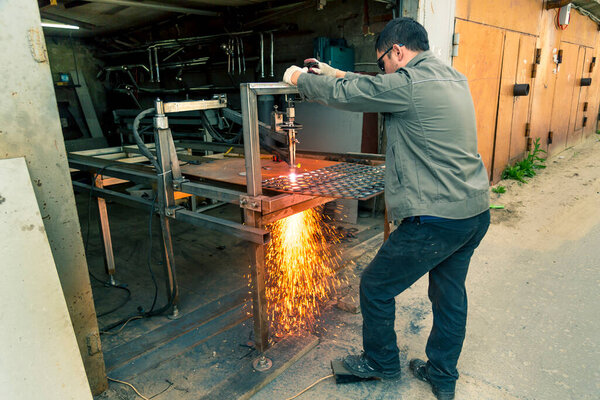 A worker cuts thick sheet steel with a plasma machine, scattering drops of molten steel in dif-ferent directions