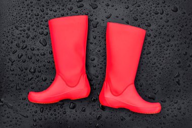 Trendy red rubber boots on black wet surface covered with raindrops. Top view, space for text.