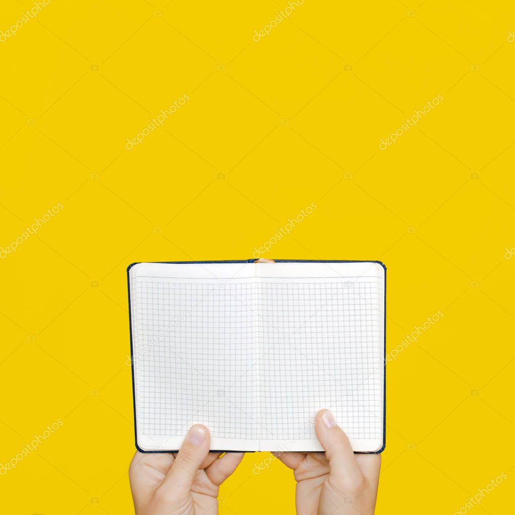 A woman's hand holds a blue closed notebook on a bright yellow background.