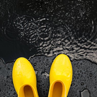 Sarı parlak gumboots standı siyah bir zemin üzerine yağmur damla su birikintisi yakınındaki kaplı. Üstten görünüm, kopya alanı