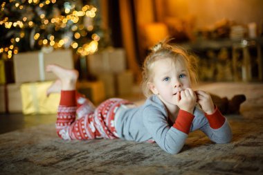 Dekore edilmiş bir Noel ağacı ve yanan çelenk arka plan üzerinde halı üzerinde büyüleyici küçük sarışın. Rahat Noel kavramı