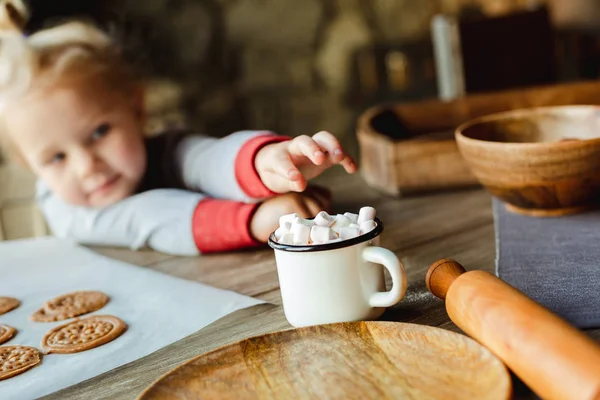 A charming little girl reaches for marshmallows in a Cup of cocoa, which stands on a table with Christmas cookie