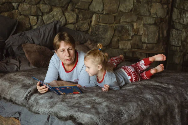 Grandma and granddaughter in Christmas pajamas reading a book, lying on the bed in the Chalet. Family Christmas concept.