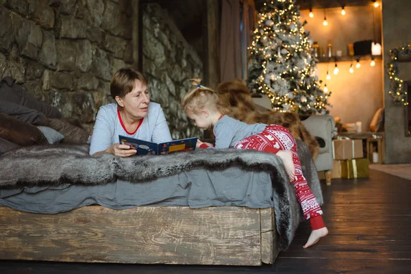 Grandma and granddaughter in Christmas pajamas reading a book, lying on the bed in the Chalet. Family Christmas concept.