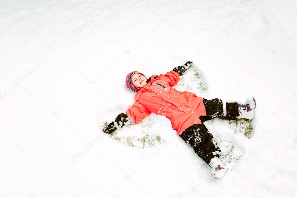 A little girl in a pink jumpsuit makes a snow angel, lying on the snow.