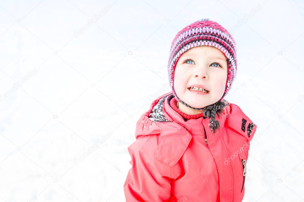 A blue-eyed little girl in a knitted hat and a pink winter jacket looks up. Snow background. Close up.