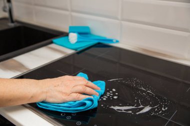 Bir kadın eli bir mavi mikrofiber bezle bir cam seramik tabak mutfakta losyonları. Arka plan vardır temizlik ürünleri.