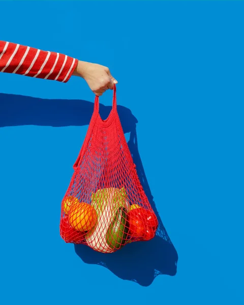 Hand with a red string bag filled with fresh vegetables and fruits from the market. The concept of zero waste and healthy eating. The style of pop a