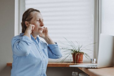 Yaşlanma önleyici ve yüz kırışıklıkları. Yaşlı bir kadın, yaşlanma karşıtı yüz kaldırma kılavuzuna egzersizlerle bakar ve onları bilgisayarın önünde tekrarlar..