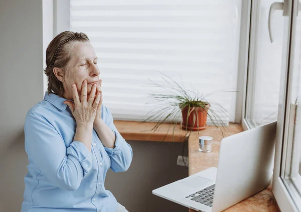 Evde online dersler var. Yaşlı beyaz bir kadın dizüstü bilgisayarın önünde yüz bakımı yapıyor. Bir salgın sırasında sağlığınıza dikkat etmek.