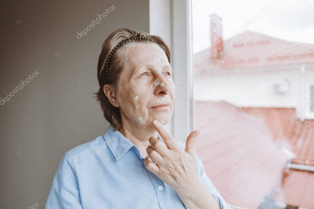 An elderly woman puts cream on her face. Home anti-aging skin care. Daily cosmetic routine.