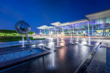 Wroclaw, Poland - June 17, 2020: Modern Wroclaw Airport terminal at dusk. Fountains in chillout zone in front of illuminated building with sky reflections on glass facade wall clipart