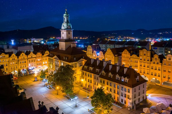 Jelenia Gora pazar meydanında yıldızlı bir gece. Güzel aydınlatılmış belediye binası, merkezdeki eski apartmanlar ve restoranlar. Dağları çevreleyen bir mesafede
