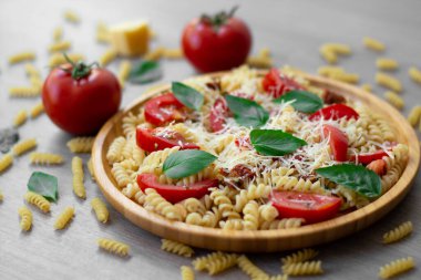 Yaz soğuk makarna salatası fusilli makarna, taze domates, güneşte kurutulmuş domates rendelenmiş parmesan peyniri ve ahşap bir tabakta taze yeşil fesleğen yaprakları domates, fesleğen, kuru makarna ve parmesan peyniri ile çevrili..