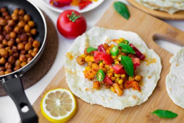 Nohutlu tortilla taco, dolmalık biber, mısır, domates, fesleğen yaprağı, çedar peynirli yeşil biber ve Monterrey Jack. Arka planda malzemeler, limonlar ve küçük bir tavada nohut var.