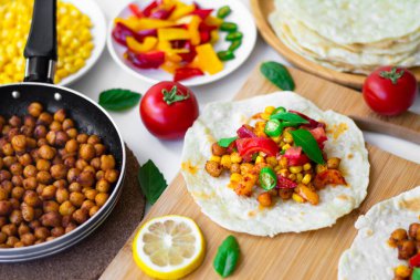 Nohutlu tortilla taco, dolmalık biber, mısır, domates, fesleğen yaprağı, çedar peynirli yeşil biber ve Monterrey Jack. Arka planda malzemeler, limonlar ve küçük bir tavada nohut var.