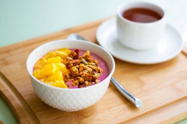 Donmuş muz ve ahudududulu granola ve taze portakalla süslenmiş pembe smoothie kasesinin sağlıklı yaz kahvaltısı. Arka tarafta siyah çay var.