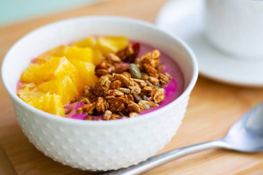 Donmuş muz ve ahudududulu granola ve taze portakalla süslenmiş pembe smoothie kasesinin sağlıklı yaz kahvaltısı. Arka tarafta siyah çay var.