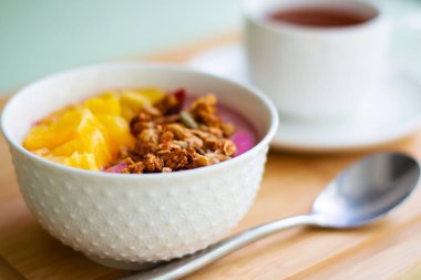 Donmuş muz ve ahudududulu granola ve taze portakalla süslenmiş pembe smoothie kasesinin sağlıklı yaz kahvaltısı. Arka tarafta siyah çay var.