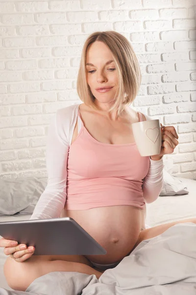 Felice donna incinta sorridente è seduto a letto e guarda tablet . — Foto Stock