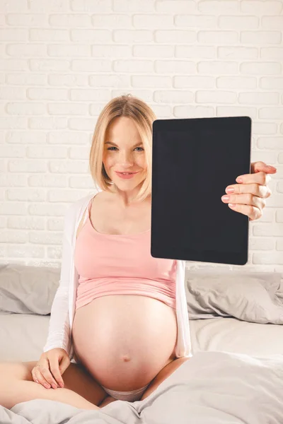 幸せな笑顔の妊娠中の女性にベッドに横し、タブレットを見て. — ストック写真