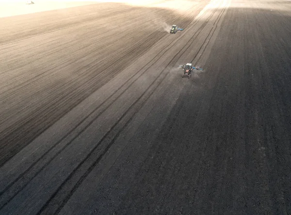 Two bright blue tractor plowing the ground against a black earth background.