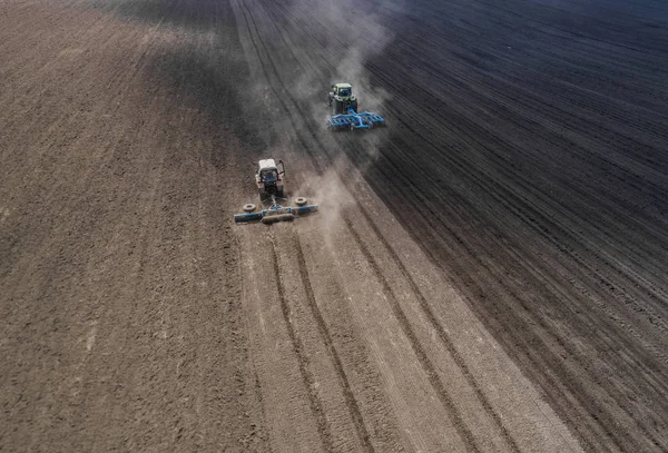 Due trattori blu brillante arare il terreno contro uno sfondo nero terra . — Foto Stock