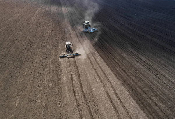 Due trattori blu brillante arare il terreno contro uno sfondo nero terra . — Foto Stock