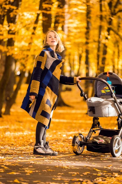 ベビーカーの若い母親は、秋の公園を歩く — ストック写真