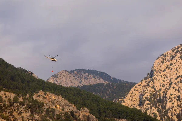 Een brand helikopter met een volledige korf van water vliegt naar het blussen van een bosbrand tegen de achtergrond van bergen, geschilderd in heldere oranje zon kleur — Stockfoto