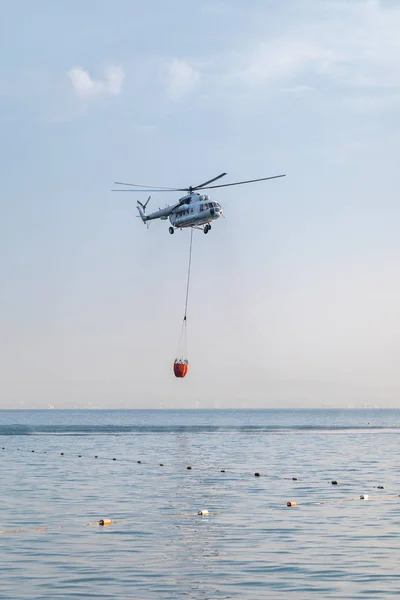 Um helicóptero com uma cesta vermelha é abaixado sobre o mar para pegar água . — Fotografia de Stock