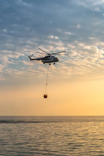 A fire helicopter collects water into the sea and flies toward the mountains to extinguish a forest fire on the background of a beautiful dawn sky — Stock Photo, Image
