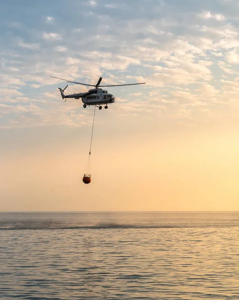 A fire helicopter collects water into the sea and flies toward the mountains to extinguish a forest fire. — Stock Photo, Image