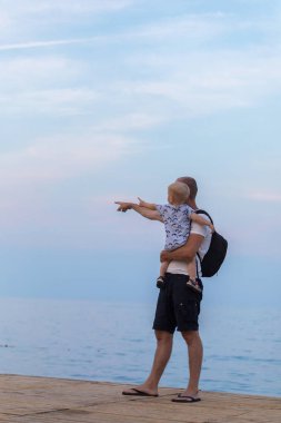 Baba oğlu onun kollarında tutar ve deniz gösterir