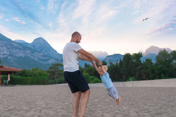 Pai e filho brincando na praia contra o pano de fundo das montanhas e uma pequena praia isolada casa unifamiliar . — Fotografia de Stock