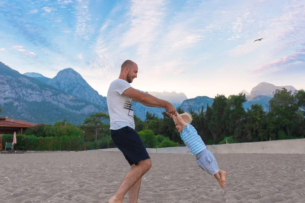 Padre e hijo jugando en la playa contra el telón de fondo de las montañas y una pequeña playa aislada casa unifamiliar . — Foto de Stock