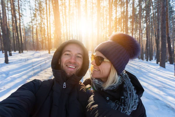Young couple in love makes selfie in the winter snow-covered forest on the background of the bright setting sun. Walks in the open air. Travel as a lifestyle, life in motion