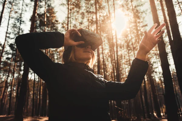 Woman in forest with virtual reality headset looking straight and trying to touch something with her hand