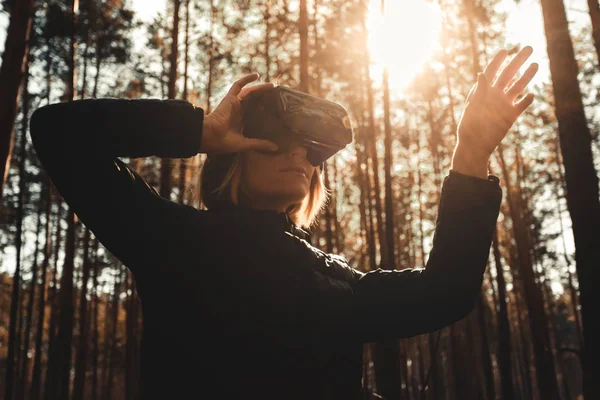 Жінка в лісі з гарнітурою віртуальної реальності дивиться прямо і намагається торкнутися чогось рукою Стокове Фото