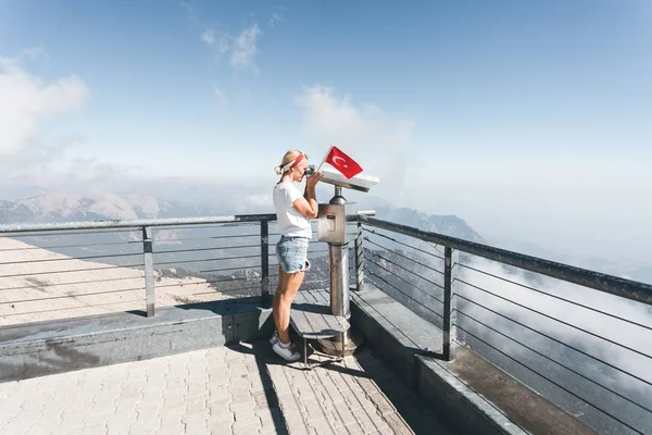Ung kvinna turist står på en utsiktsplattform av Tahtali Mountain i Kemer med en röd kalkon flagga och ser i kikare, Turkiet — Stockfoto