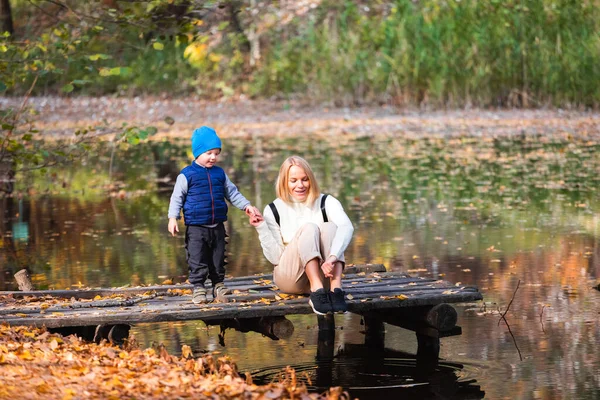 Happy young mother and her son spending time in the autumn park near the pond.