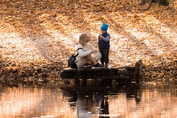 Mãe e filho estão sentados em um cais de madeira perto do lago no parque de outono — Fotografia de Stock