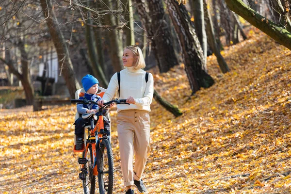 Felice madre conduce una bicicletta con un bambino legato nella parte posteriore nel parco autunnale. — Foto Stock