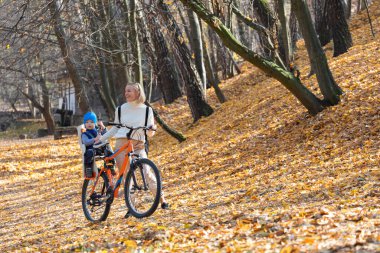 Happy mother leads a bicycle with a child strapped in the back in the autumn park. clipart