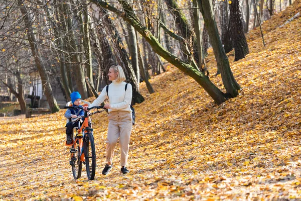 Šťastná matka vede kolo s dítětem připoutaným vzadu v podzimním parku. — Stock fotografie