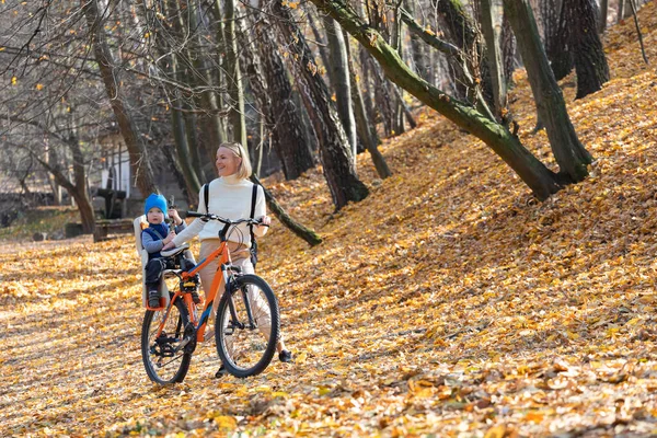 Mãe feliz leva uma bicicleta com uma criança amarrada nas costas no parque de outono. — Fotografia de Stock