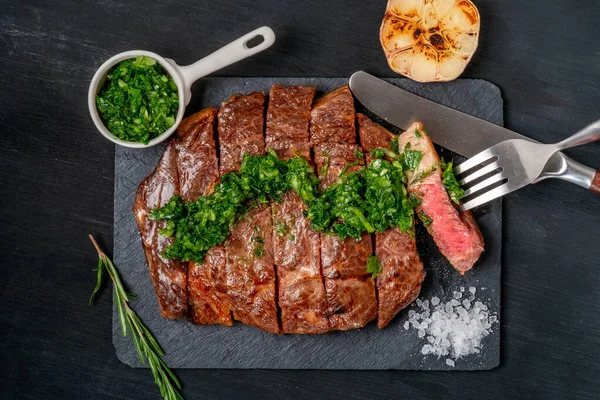 Ready to eat black Angus beef rib eye steak sliced with herbs, garlic and sauce on slate Board. Ready meal for dinner on the background of dark black wood. Top view with copy space